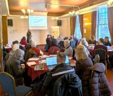 Photograph of a group of people sat around tables talking. With a presentation and speaker in the background