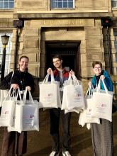 photograph of three people holding up bags smiling into the camera, with a historic building in the background