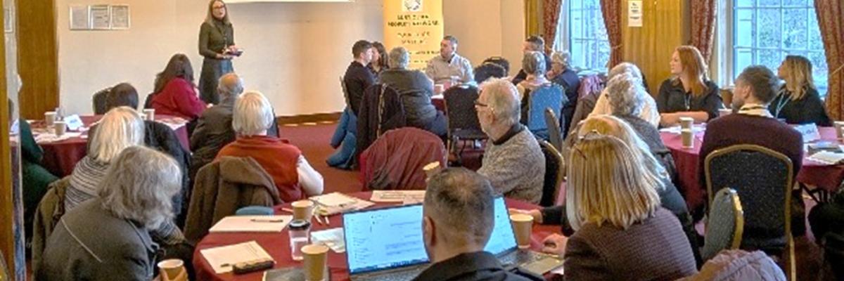 Photograph of a group of people sat around tables talking. With a presentation and speaker in the background