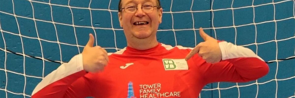 Photo of man smiling into the camera with a thumbs up gesture wearing a red shirt. Background includes a goalkeepers football net.
